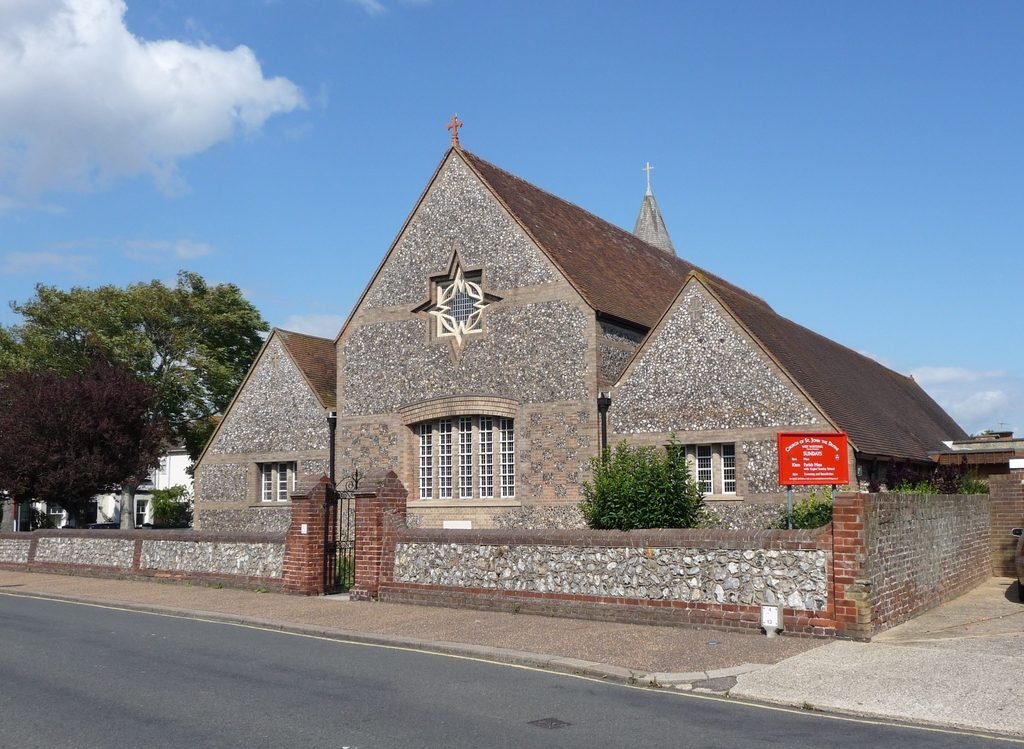 St John the Divine seen from Ripley Road, its flint walls and rose window bright under a blue sky.