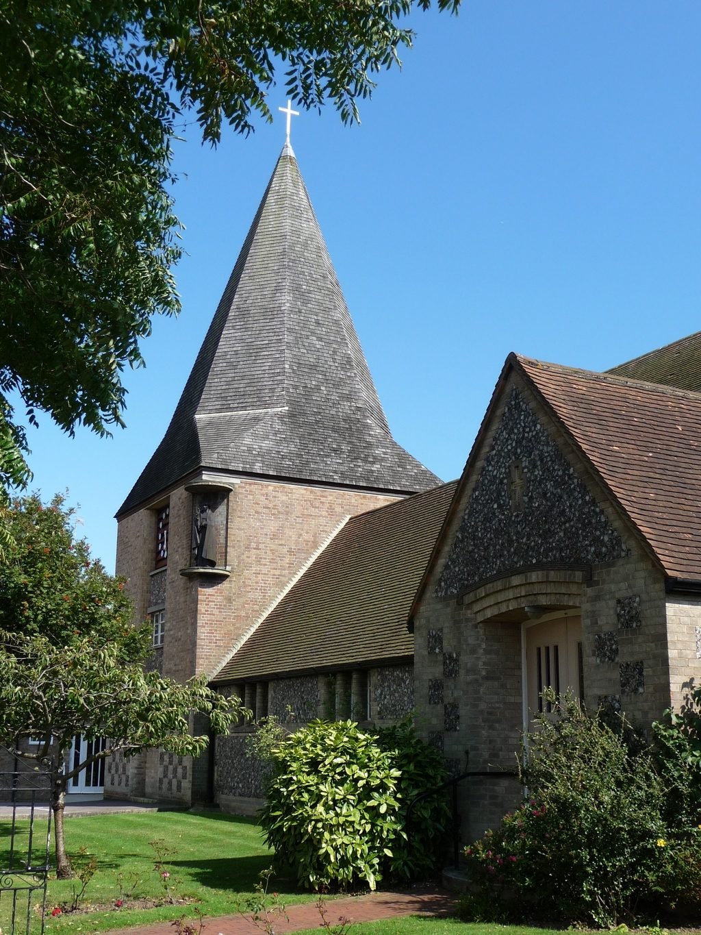 The spire of St John the Divine rising above the tiled roof and porch, framed by summer trees.