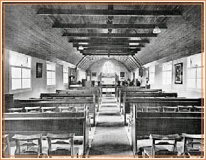 The interior of the mission room, with rows of simple wooden benches beneath a boarded roof.